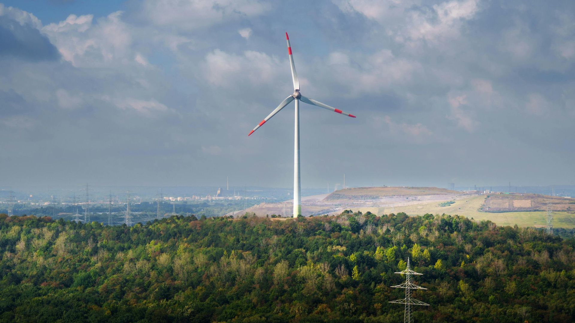 A single wind turbine with trees in the foreground and buildings and electricity pylons in the background
