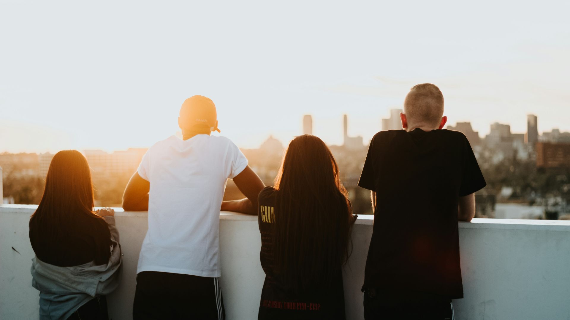 A group of four young people looking out from a balcony towards a sun setting on a city skyline. Photo credit: Devin Avery on unsplash